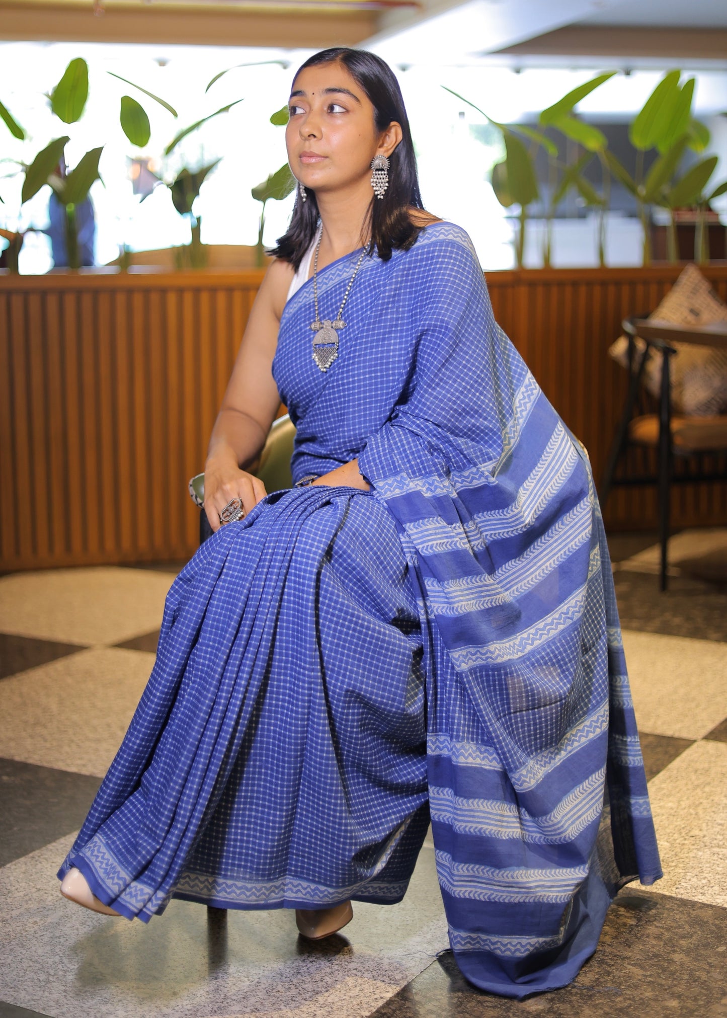 Woman in a blue saree sitting indoors with plants in the background
