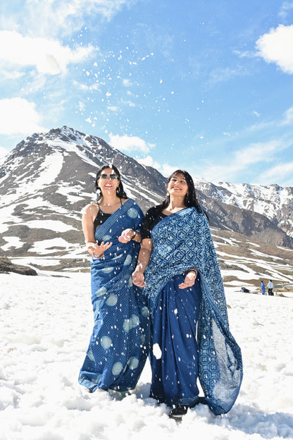 Two women wearing blue patterned sarees standing in snow with snow-capped mountains and a blue sky in the background.