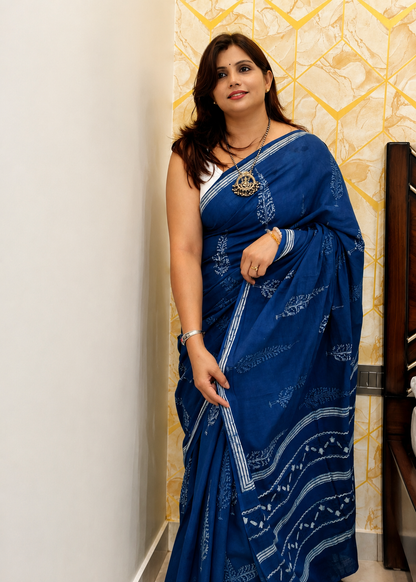 A woman wearing a royal blue saree with white bandhani patterns, standing in an indoor setting with a patterned beige wall behind her.