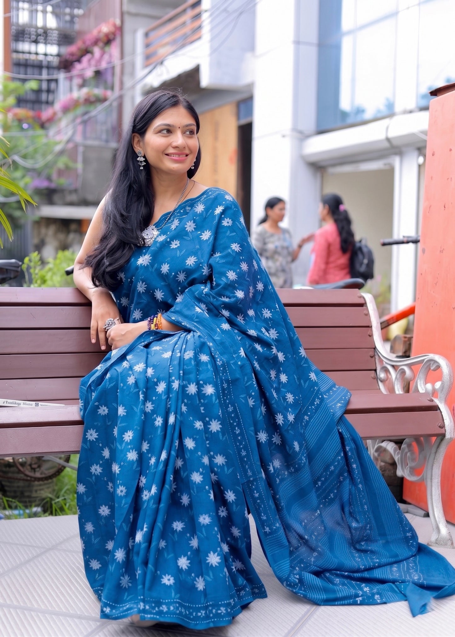 Woman in a blue saree sitting on a bench with a building and people in the background