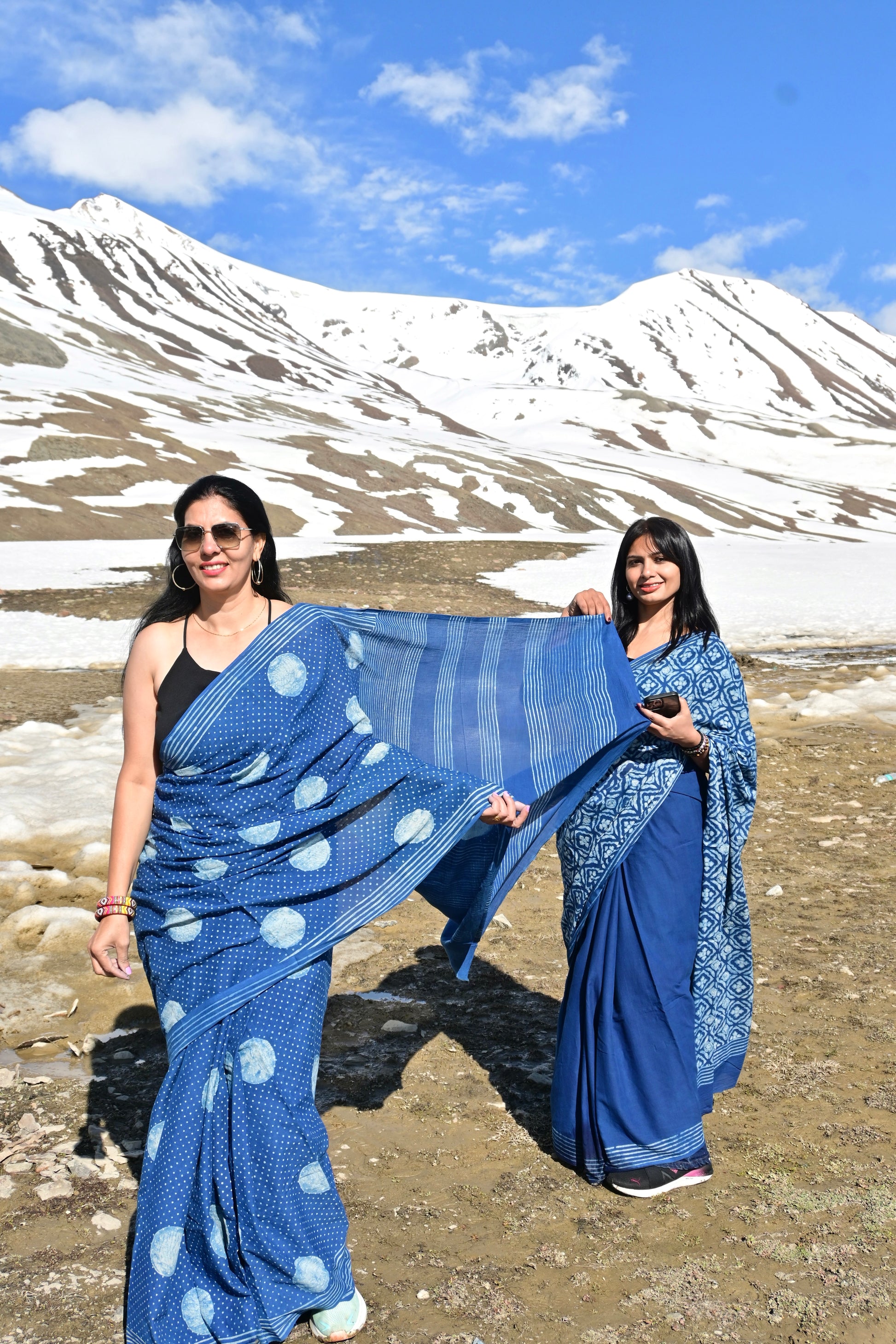 Two women wearing blue patterned sarees posing outdoors in a mountain landscape with snow and blue sky in the background.
