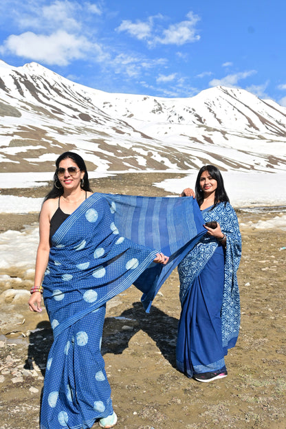Two women wearing blue patterned sarees posing outdoors in a mountain landscape with snow and blue sky in the background.