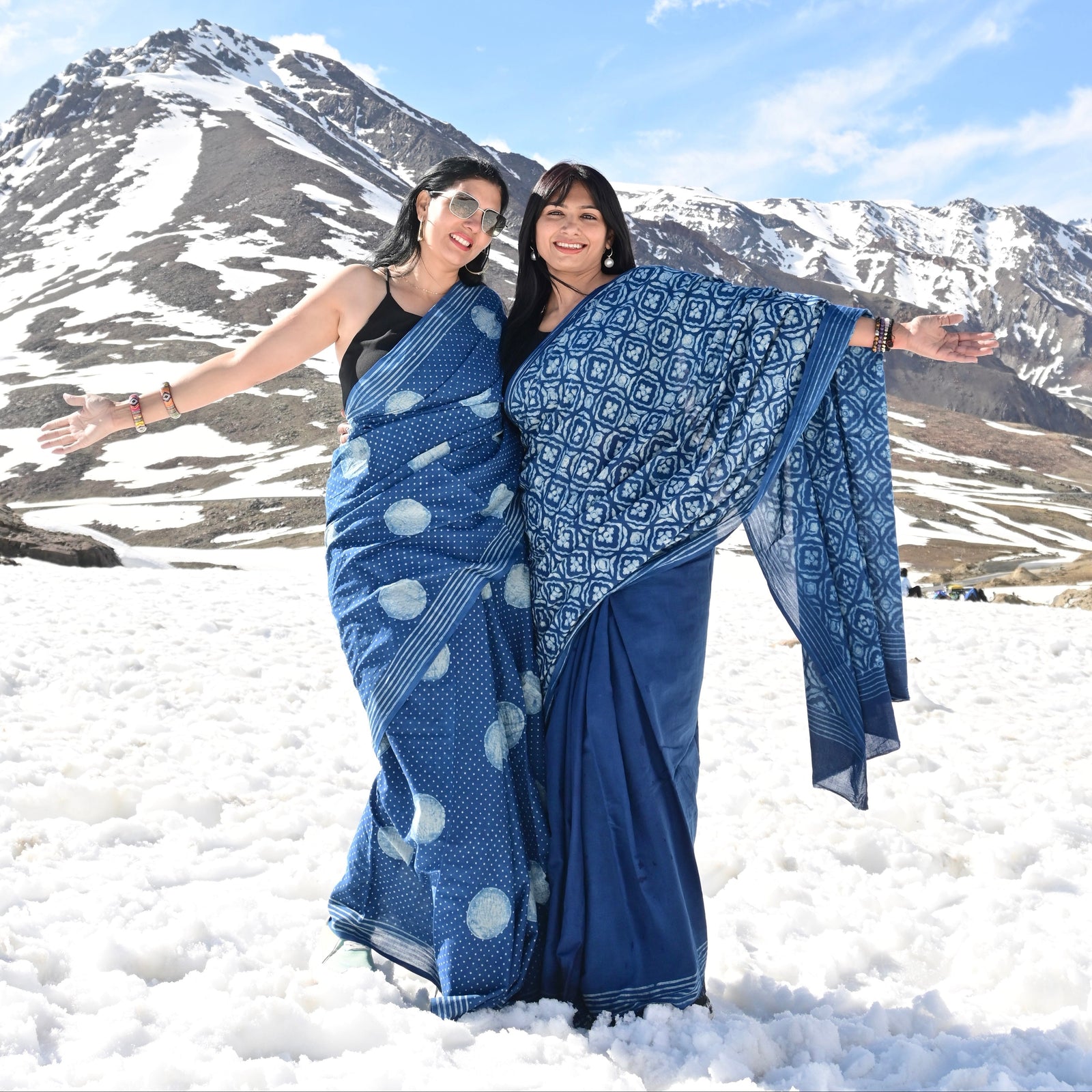 Two women posing in blue patterned sarees in a snowy mountain landscape under a blue sky.