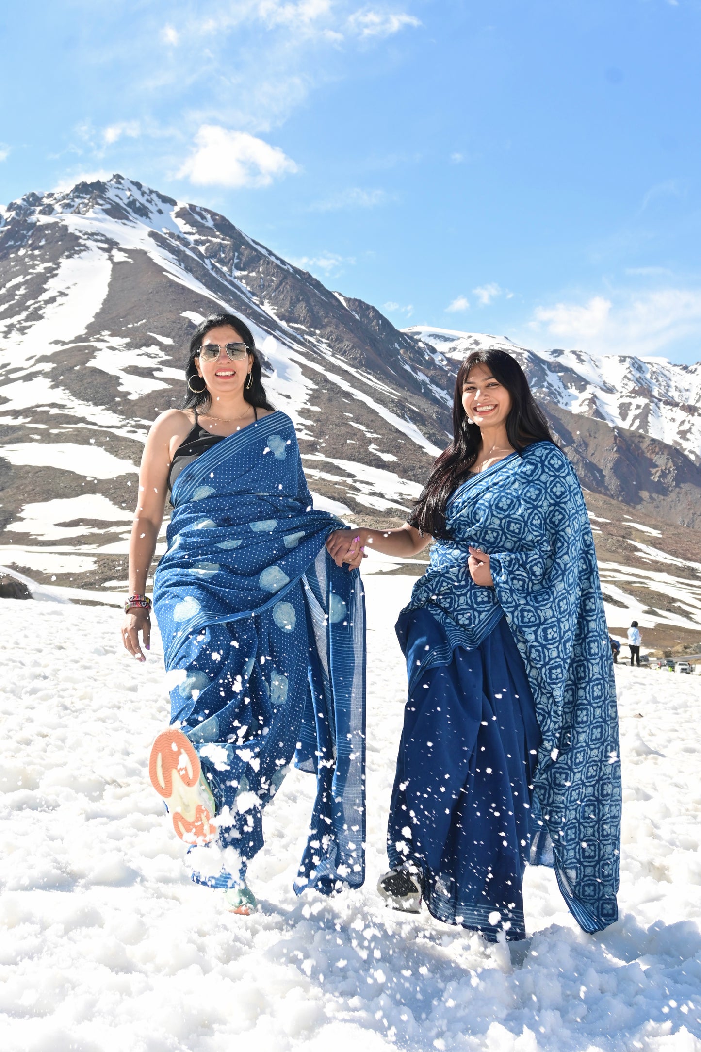 Two women wearing blue patterned sarees walk hand in hand through deep snow, with a majestic snow-capped mountain range and blue sky in the background.