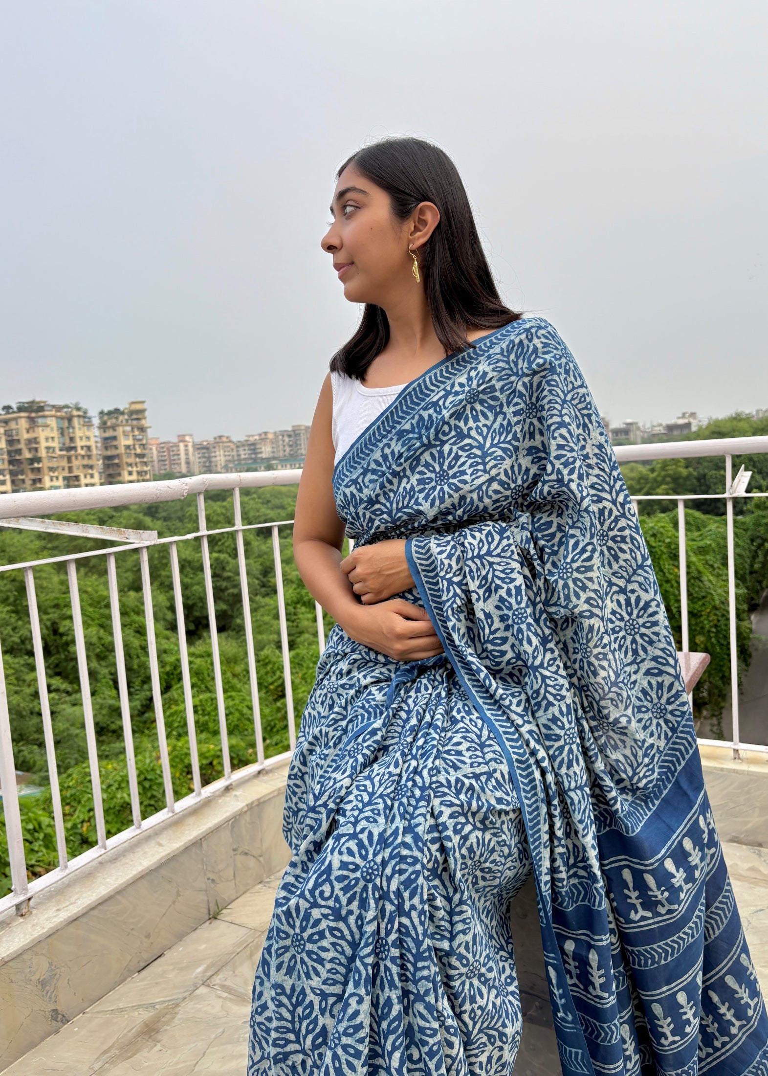 Woman wearing a blue saree with a white pattern on a balcony with greenery in the background