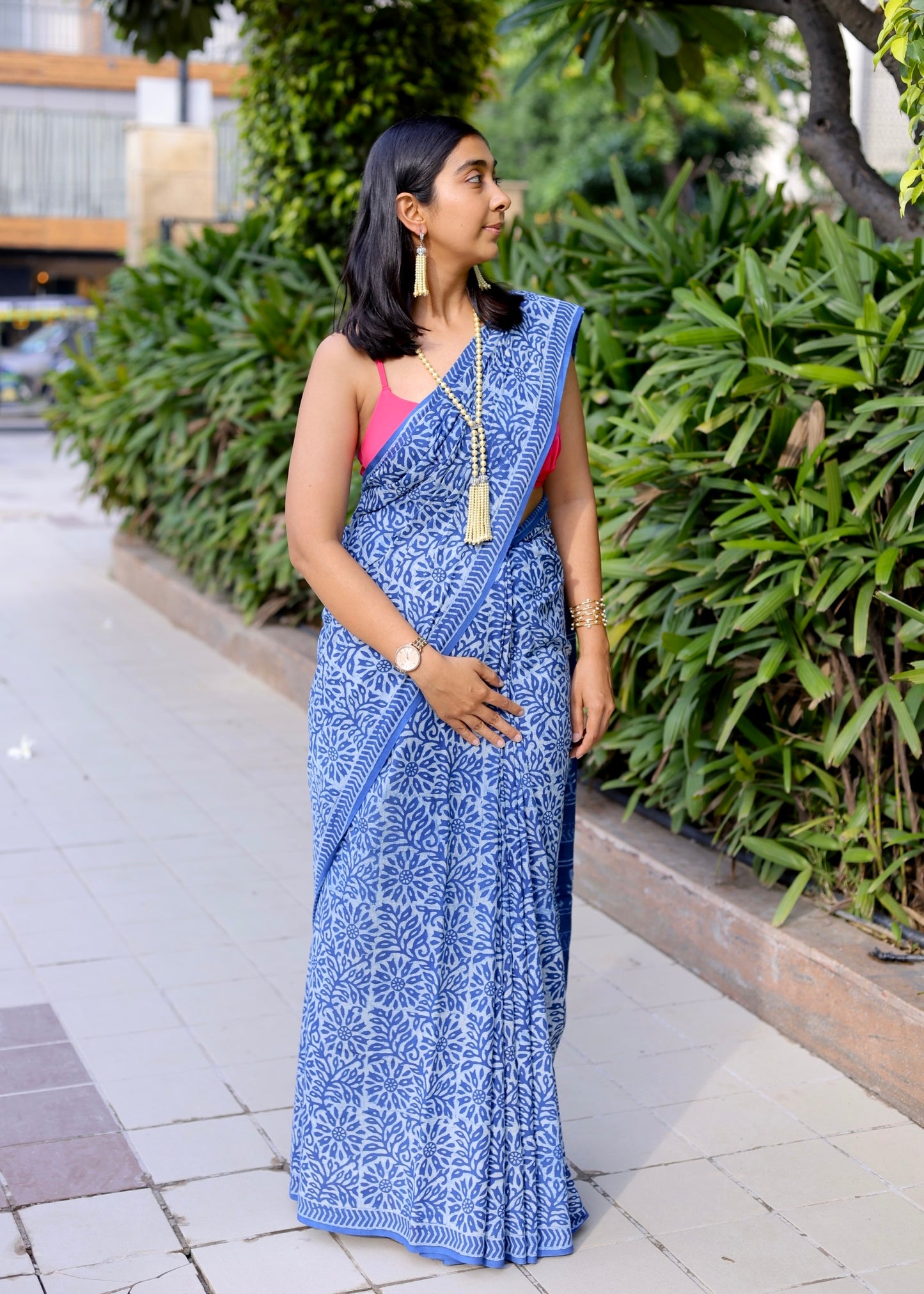 Woman wearing a blue saree with a pink blouse standing outdoors near greenery.