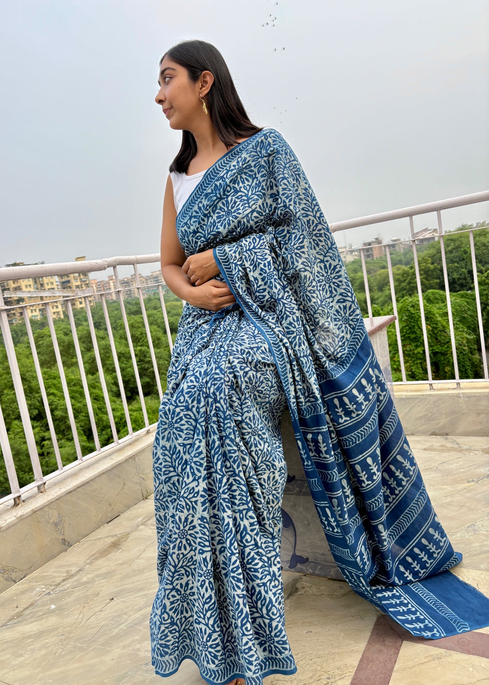 Woman wearing a blue patterned saree on a rooftop with greenery in the background