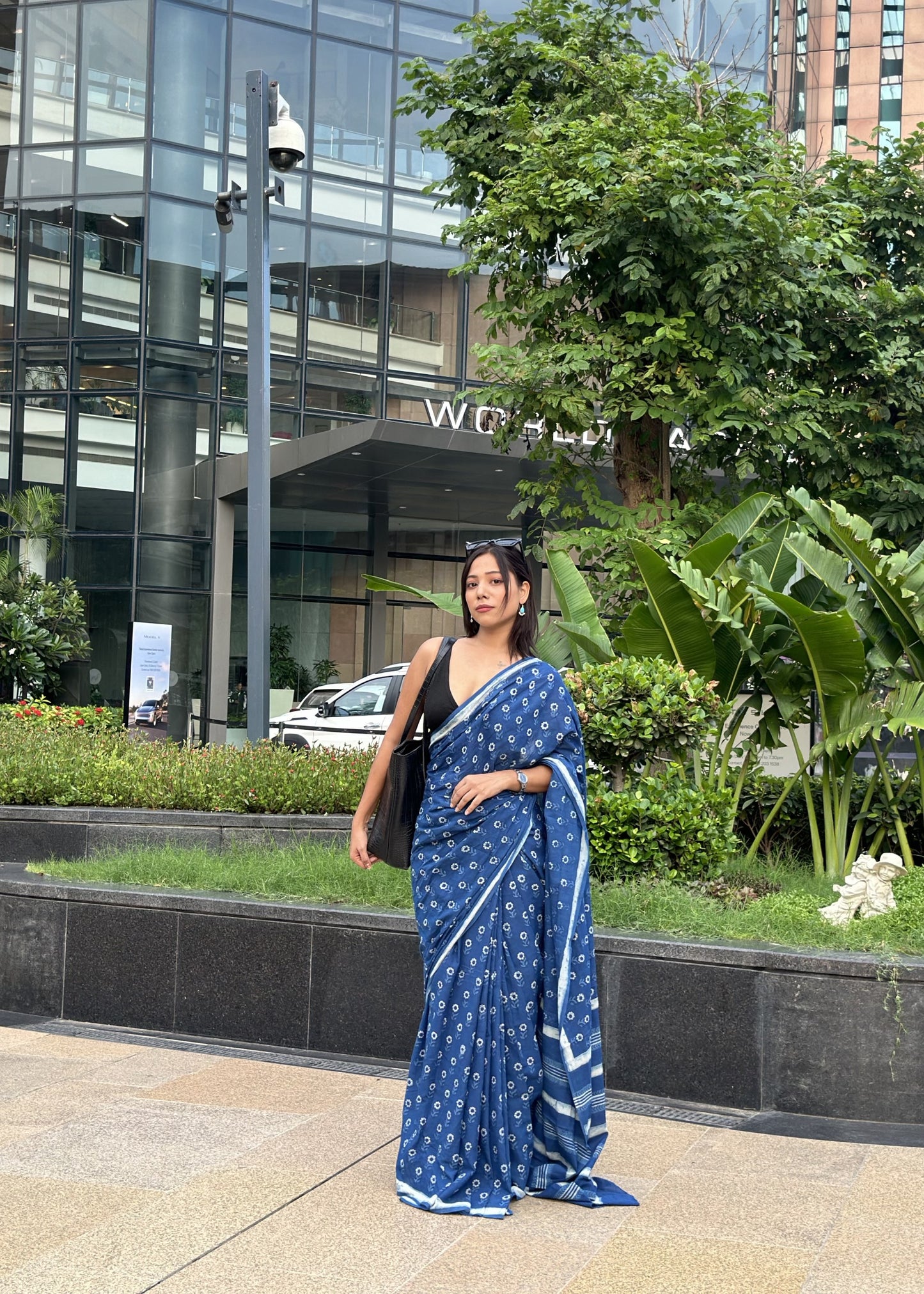 Woman in a blue saree standing in front of a modern building with greenery.
