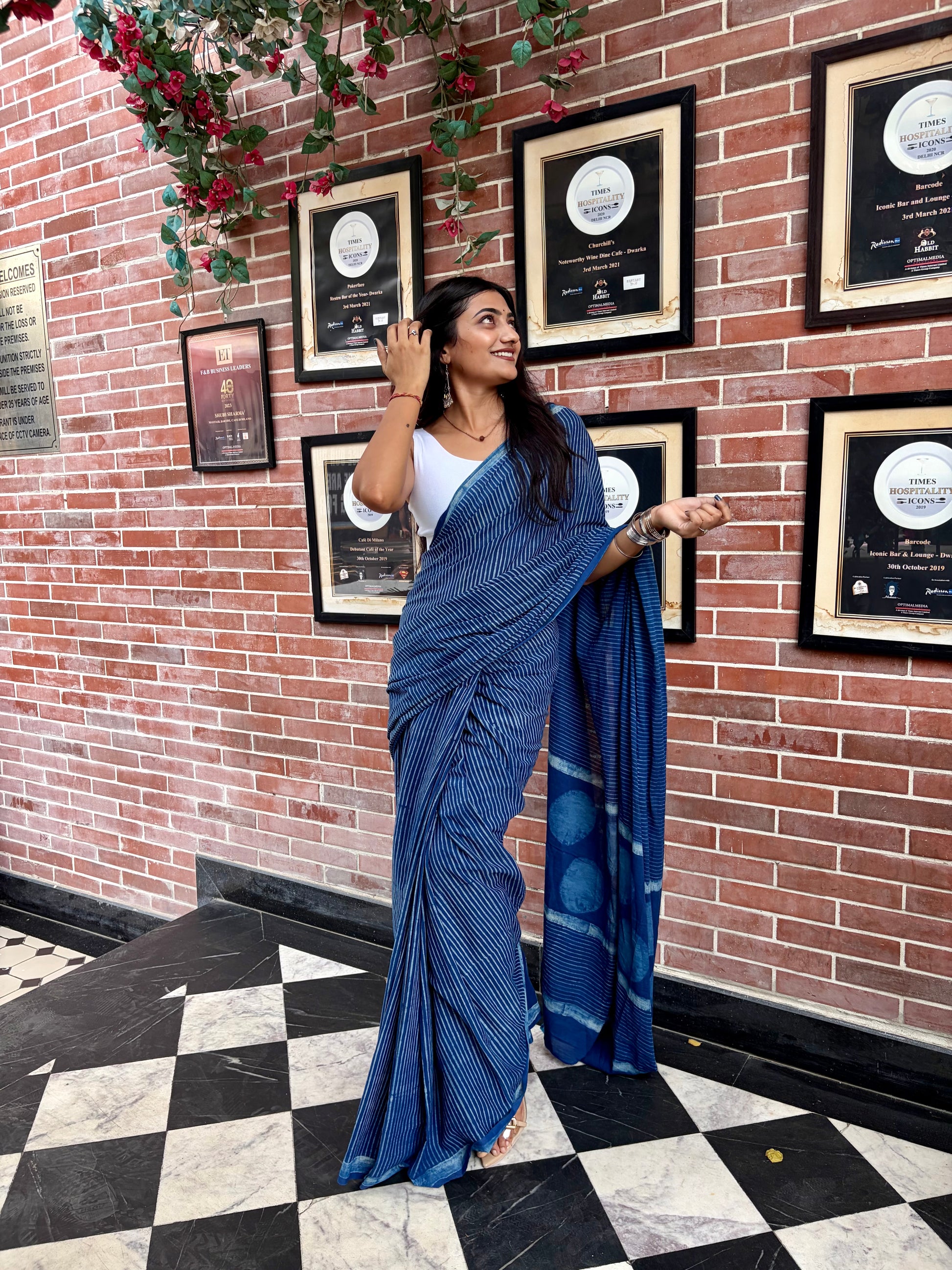 A woman wearing a blue Indigo saree and white blouse stands in front of a red brick wall with framed certificates, posing with the saree draped over one shoulder.