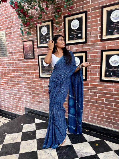 A woman wearing a blue Indigo saree and white blouse stands in front of a red brick wall with framed certificates, posing with the saree draped over one shoulder.