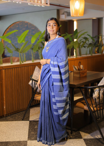 Woman in a blue saree standing in a restaurant setting with plants and tables in the background.