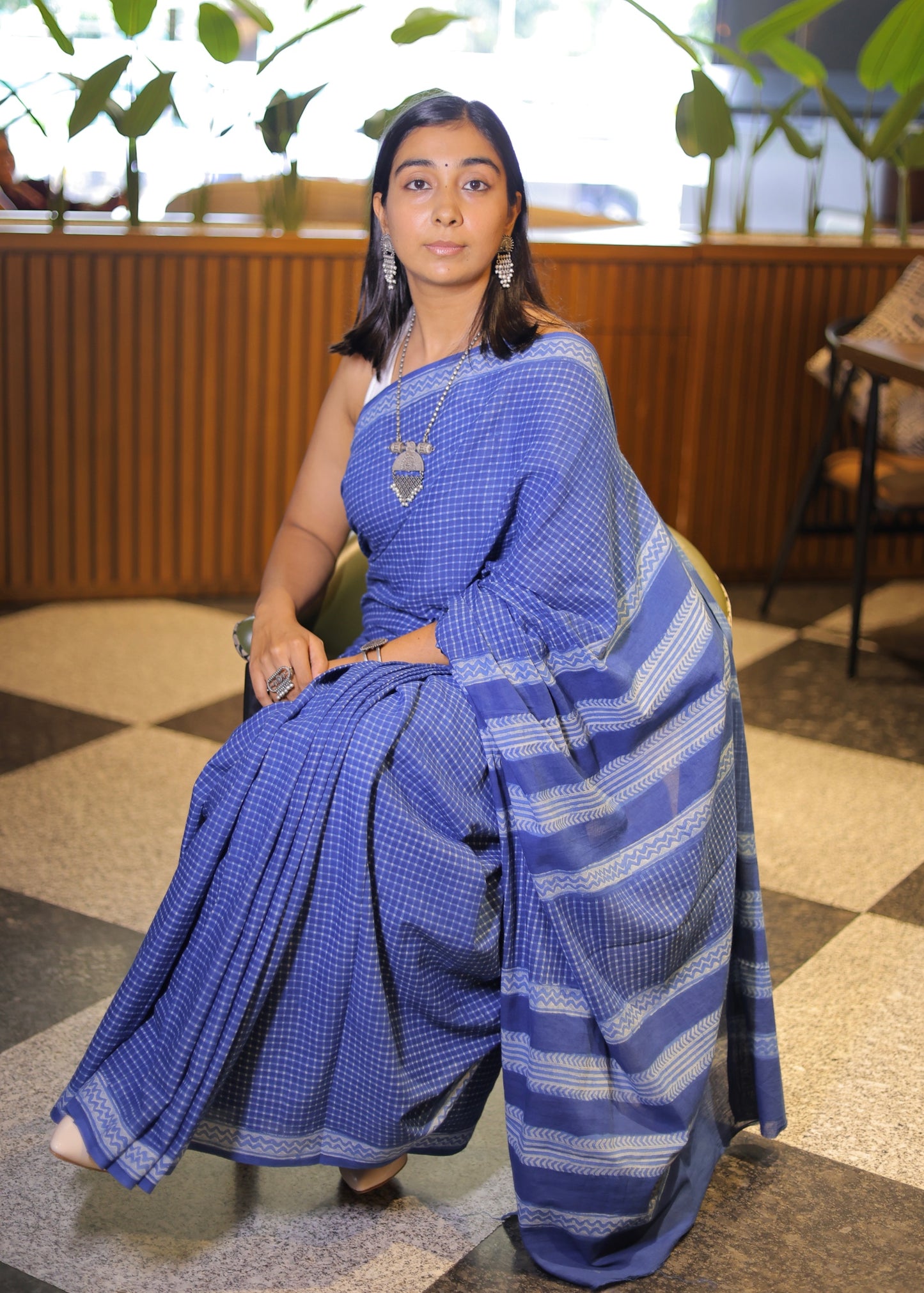 Woman in a blue saree sitting indoors with plants in the background