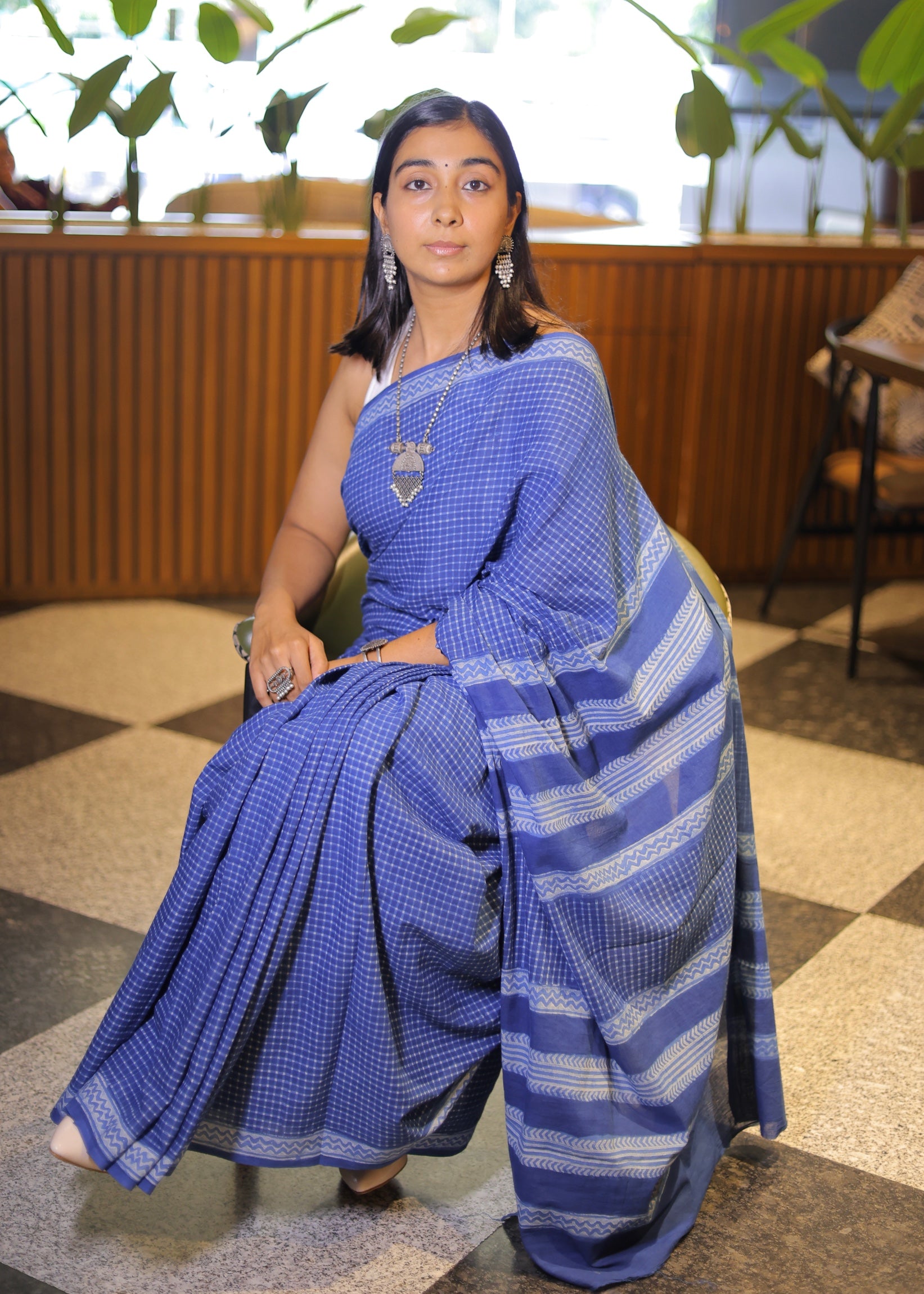 Woman in a blue saree sitting indoors with plants in the background