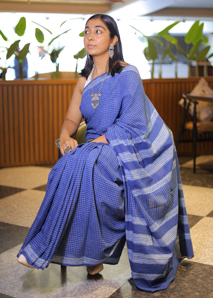 Woman in a blue saree sitting indoors with plants in the background